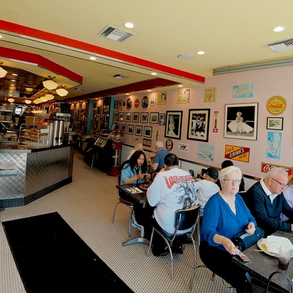 Diners eat at tables in a retro-themed restaurant with pink walls adorned with vintage signs.
