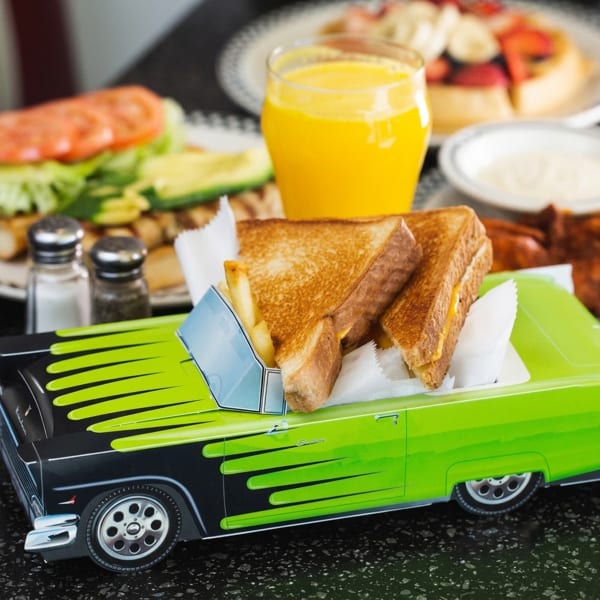 Grilled cheese and fries served in a green toy car, with orange juice and breakfast in the background.
