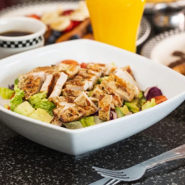 Grilled chicken salad with lettuce, avocado, and tomatoes in a white bowl.