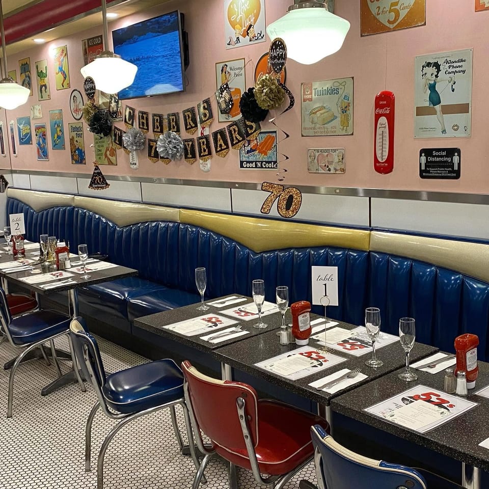 A retro diner interior with blue booths, black tables set with champagne flutes, and vintage decor.