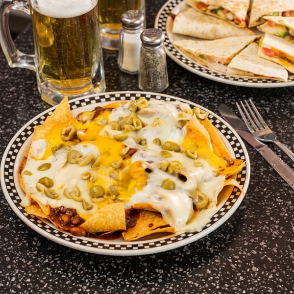 Nachos with melted cheese and green olives, beer mugs, and quesadillas on a black speckled table.