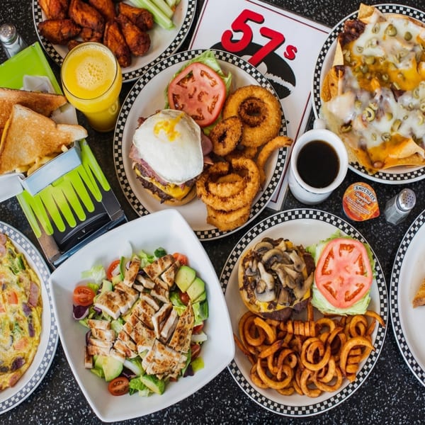 A diner table laden with various dishes, including burgers, onion rings, nachos, salad, and wings.