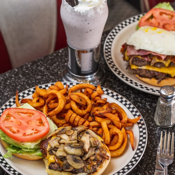 Two burgers with fries and a milkshake with whipped cream, Oreos, and a cherry on a diner table.